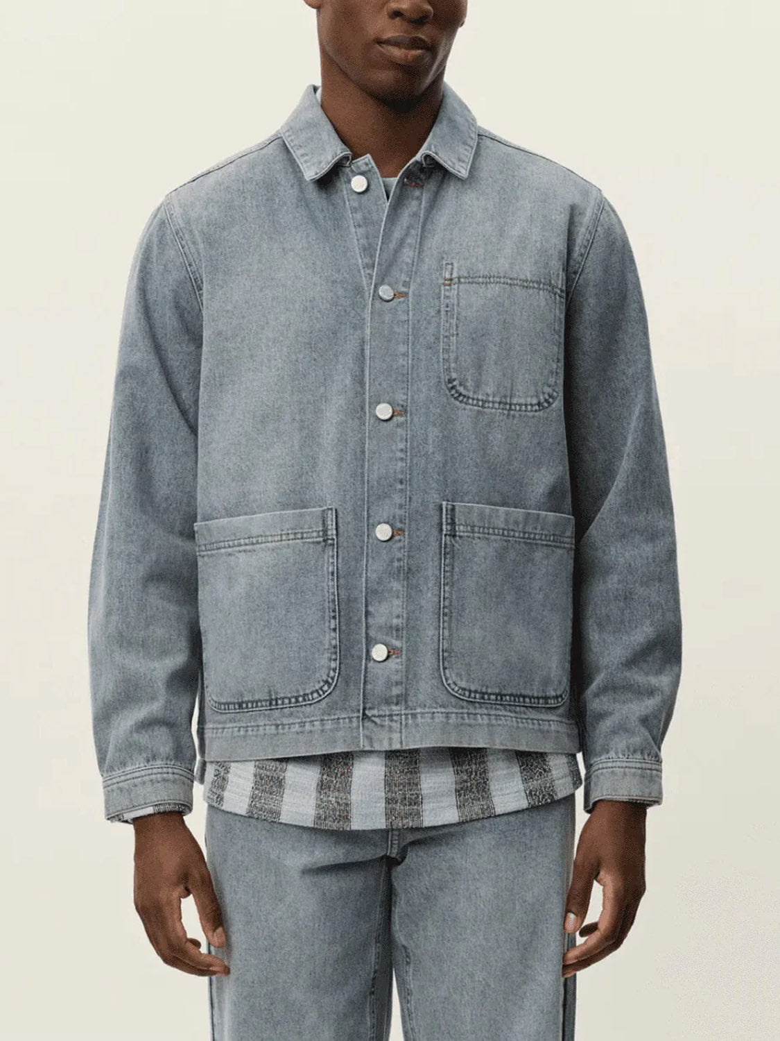 A person stands against a plain light background wearing the Le Deux Layton Denim Overshirt in light blue over a striped shirt, paired with matching denim pants and featuring large front pockets.