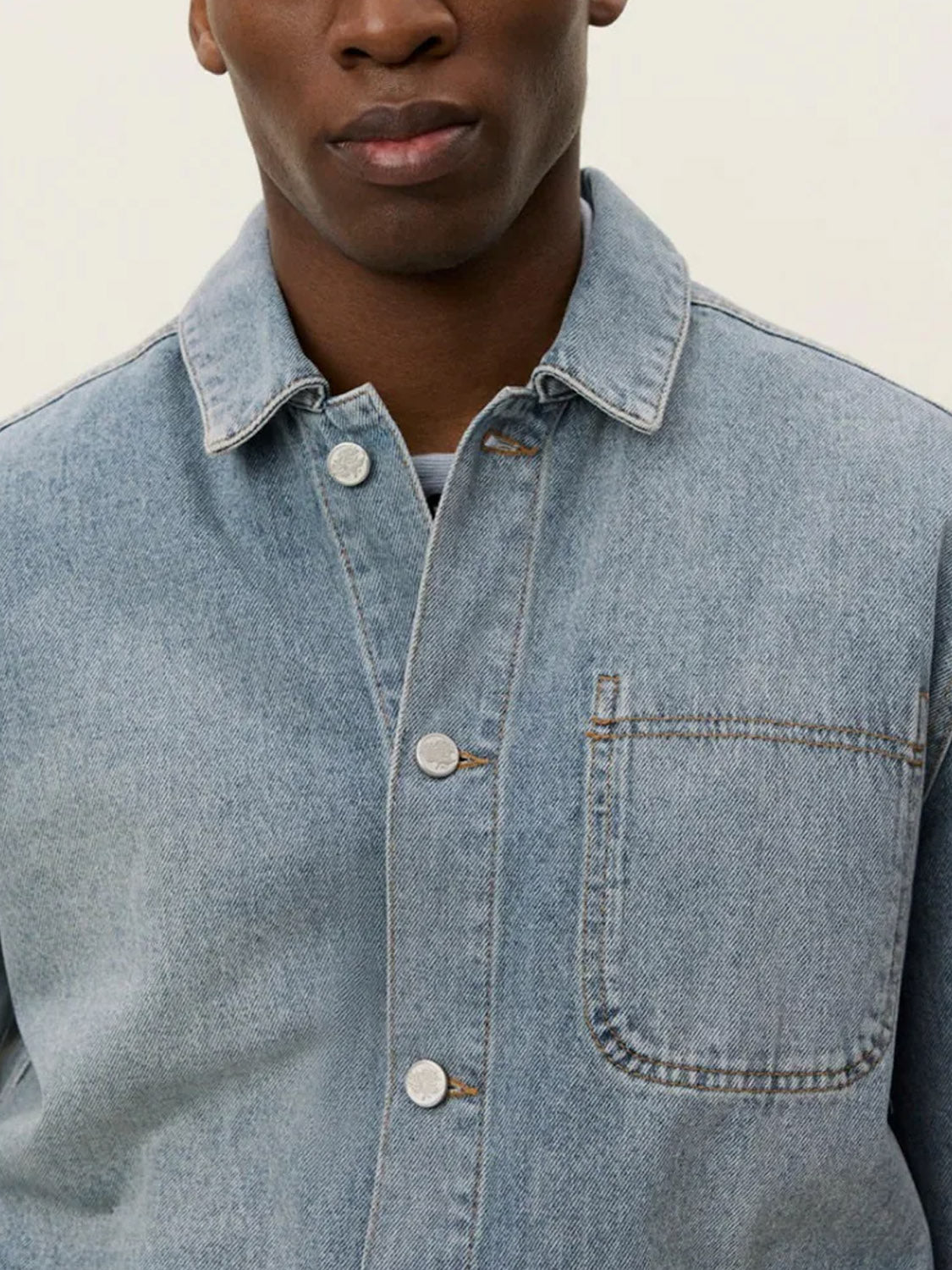 A person wearing the Le Deux Layton Denim Overshirt in light blue, regular fit, with a chest pocket and buttoned up, stands against a plain light background. Only their lower face and upper body are visible.