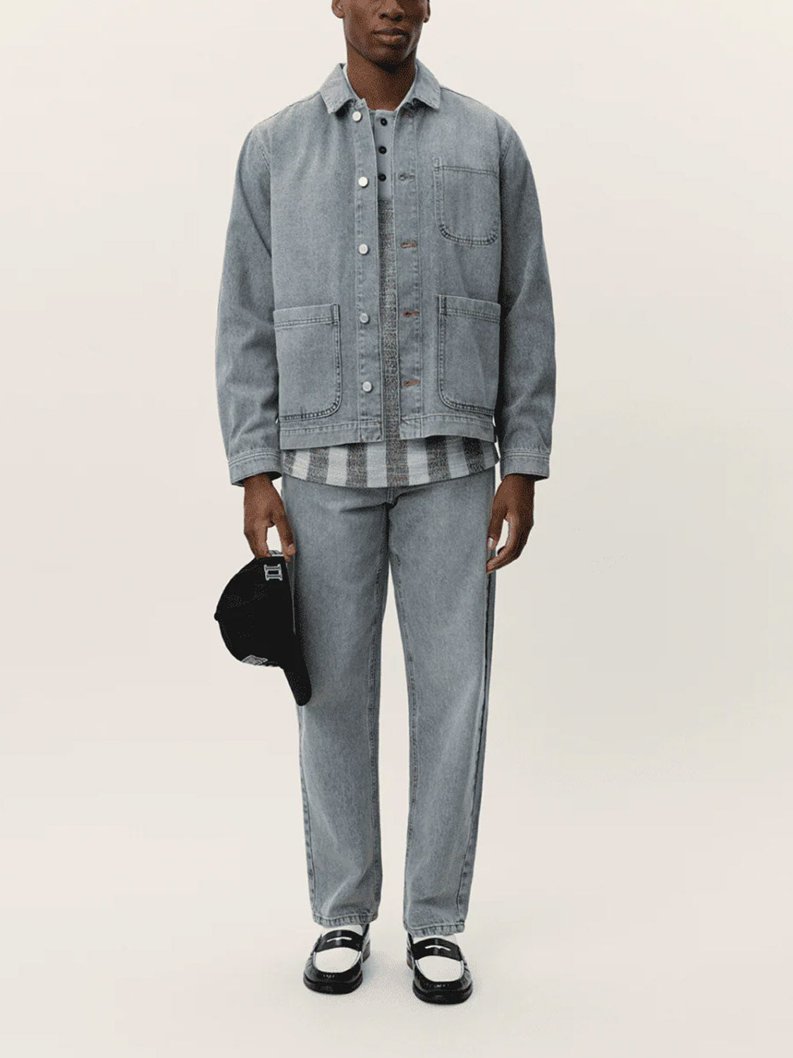 Dressed in the Le Deux Layton Denim Overshirt and matching jeans, this person holds a black cap. A gray striped shirt and black-and-white loafers complete the look against a simple, light background.
