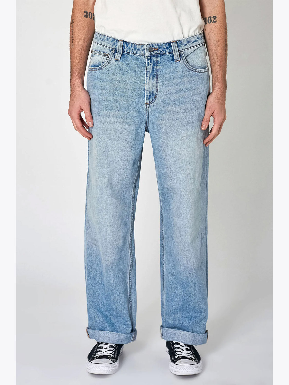 A person wears Rollas Lazy Boy Original Stone Denim jeans with rolled cuffs, a white T-shirt, and black-and-white sneakers. Standing facing forward, their relaxed arms at their sides display forearm tattoos.
