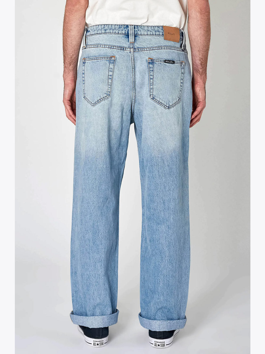Someone stands with their back to the camera in light blue Lazy Boy Original Stone Denim jeans by Rollas, featuring a wide fit, rolled cuffs, two back pockets, a brown waistband patch, and white sneakers against a plain background.