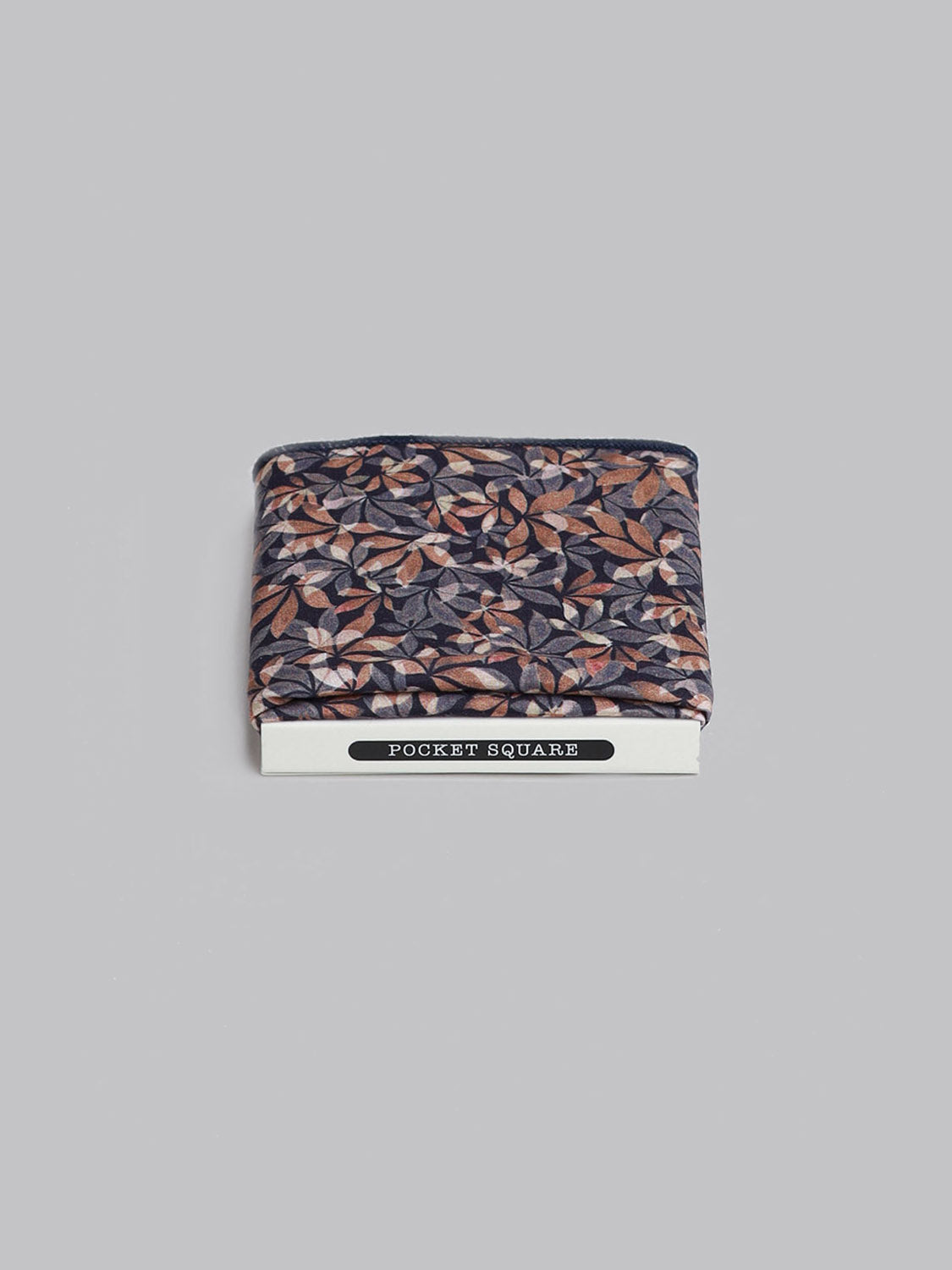 A folded Liberty™ Leaves Pocket Square by ID Menswear, featuring a brown, grey, and beige floral print on Tana Lawn cotton, is shown on a white card marked POCKET SQUARE against a plain light grey background.