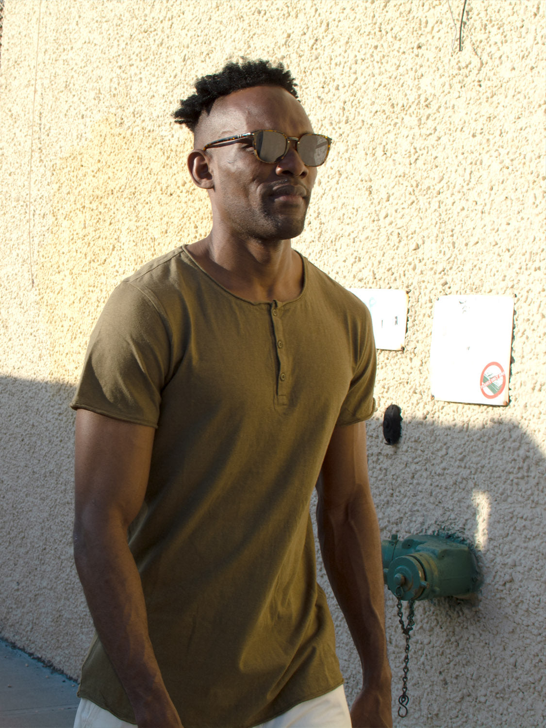 A man with short curly hair and sunglasses walks along a sunlit textured wall, wearing the Legend Hemp Cotton Rolled Neck Henley by ID Menswear. Signs and a green fixture appear in the background.