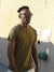 A man with short curly hair and sunglasses walks along a sunlit textured wall, wearing the Legend Hemp Cotton Rolled Neck Henley by ID Menswear. Signs and a green fixture appear in the background.