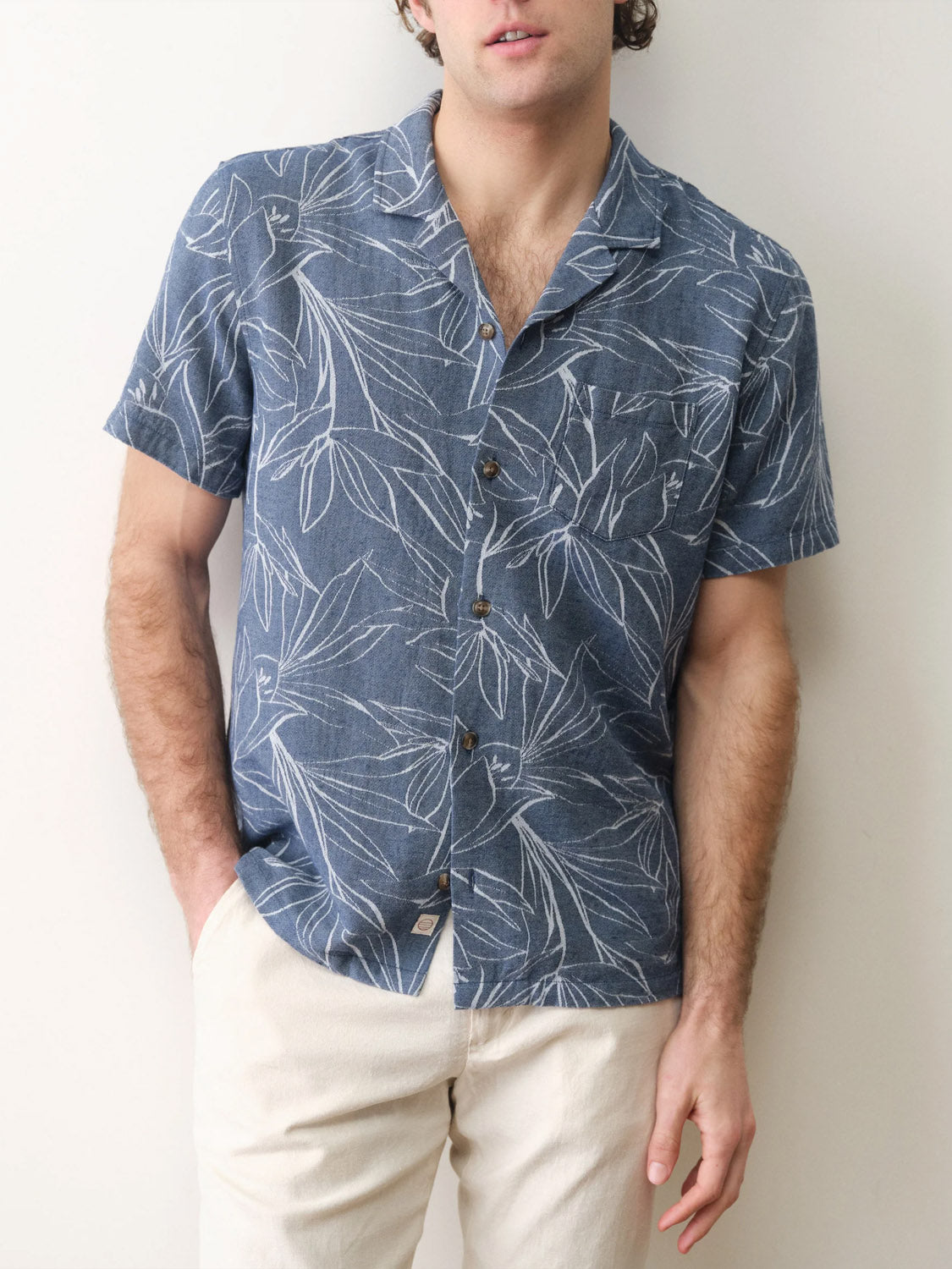A man wearing the Marine Layer Leo Jacquard Resort Shirt—a short-sleeve, blue shirt with a woven leaf design—and light-colored pants stands against a plain light wall, one hand in his pocket.