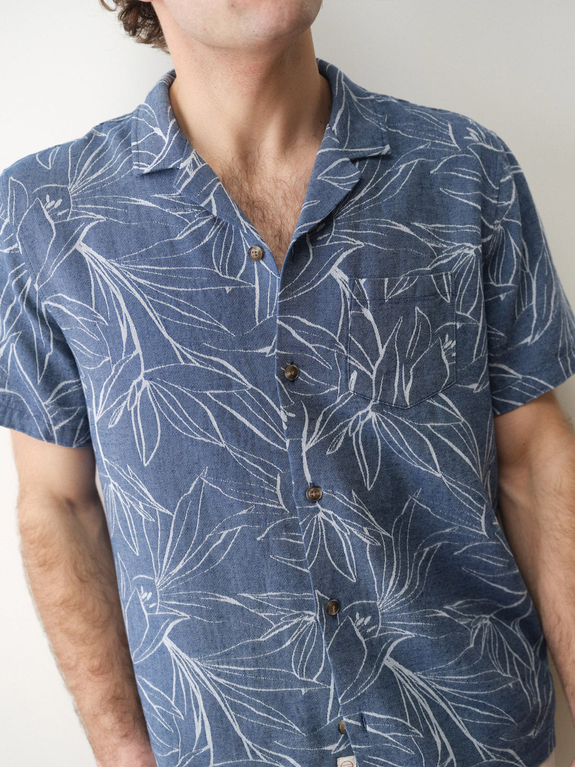 A person wears the Marine Layer Leo Jacquard Resort Shirt, a blue short-sleeve top with a woven leaf and white flower pattern, standing against a plain light background. The face is partially out of frame.