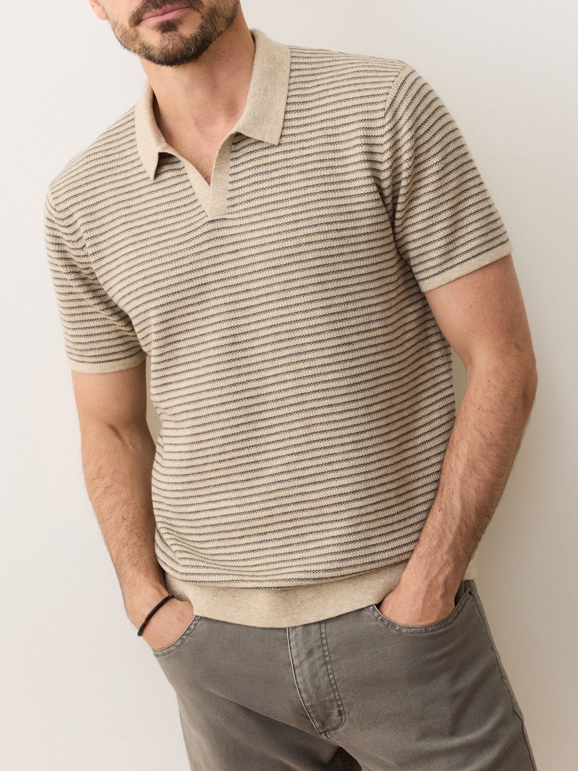 A man stands with his hands in his pockets against a plain light-colored wall, wearing the Marine Layer Liam Sweater Polo— a short-sleeve, beige and brown striped Merino wool top—paired with gray pants.