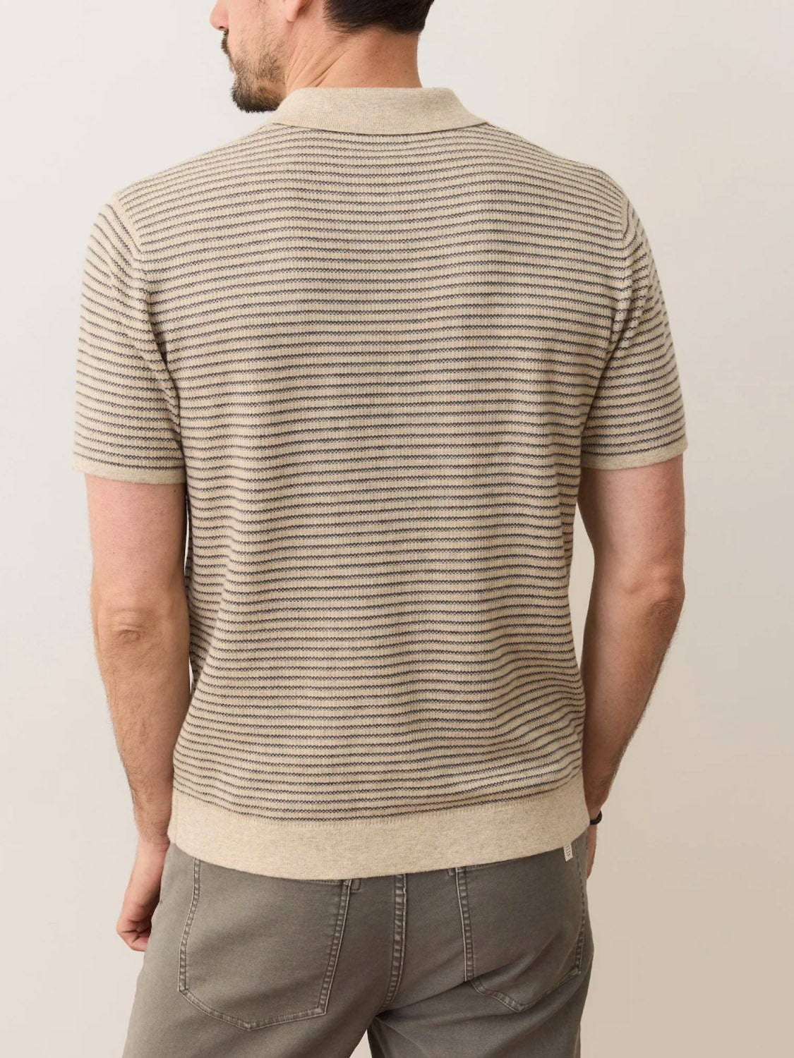 A man is seen from behind wearing the Marine Layer Liam Sweater Polo in beige and grey stripes with grey pants, standing against a plain light background.