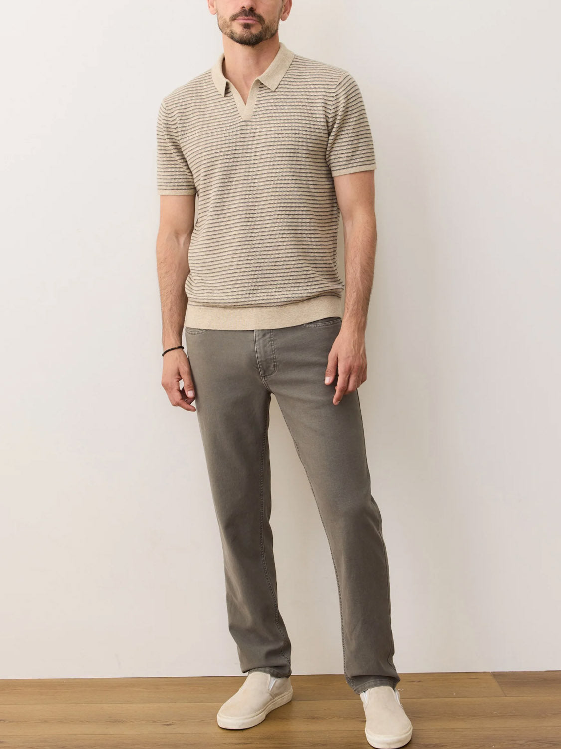 A man with a short beard stands relaxed with one hand in his pocket, wearing the Marine Layer Liam Sweater Polo, gray jeans, and light-colored slip-on shoes against a plain wall.