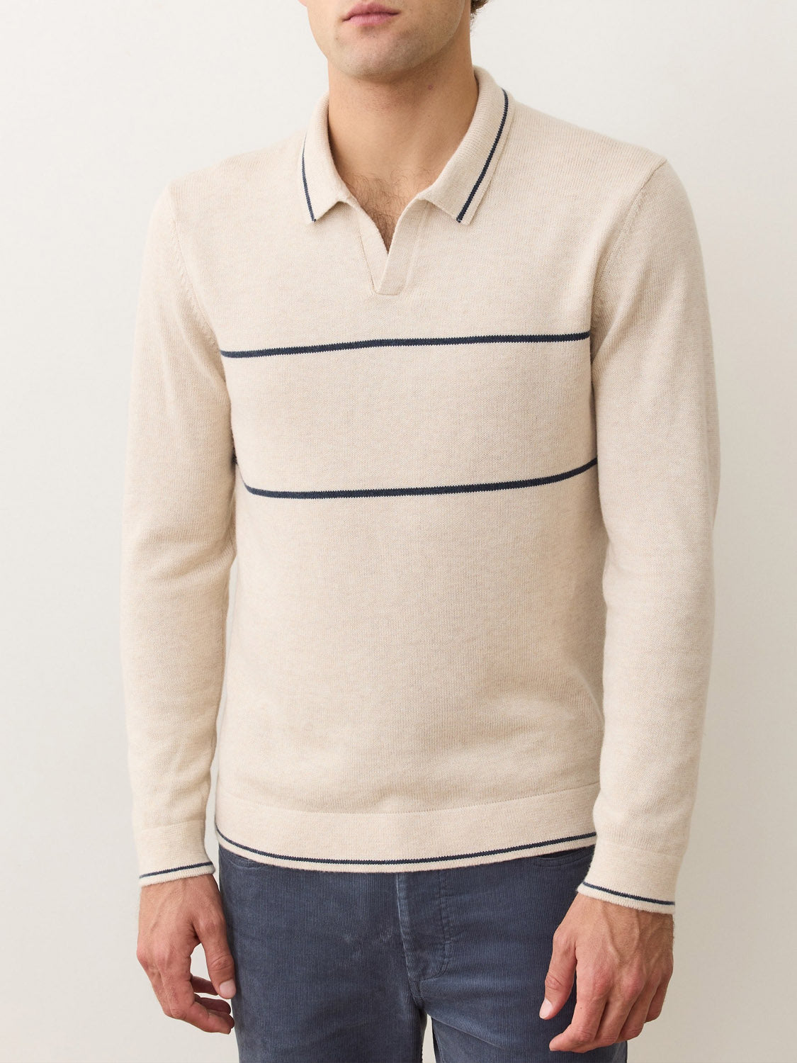 A man wearing the Marine Layer Liam Long Sleeve Sweater Polo in beige with a Johnny collar and navy stripes across the chest, paired with dark blue jeans.