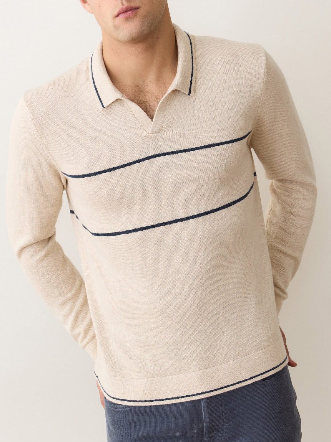 A man stands against a light background with his hands behind his back, wearing the Marine Layer Liam Long Sleeve Sweater Polo—a beige merino wool knit shirt with a Johnny collar and thin navy blue horizontal stripes.