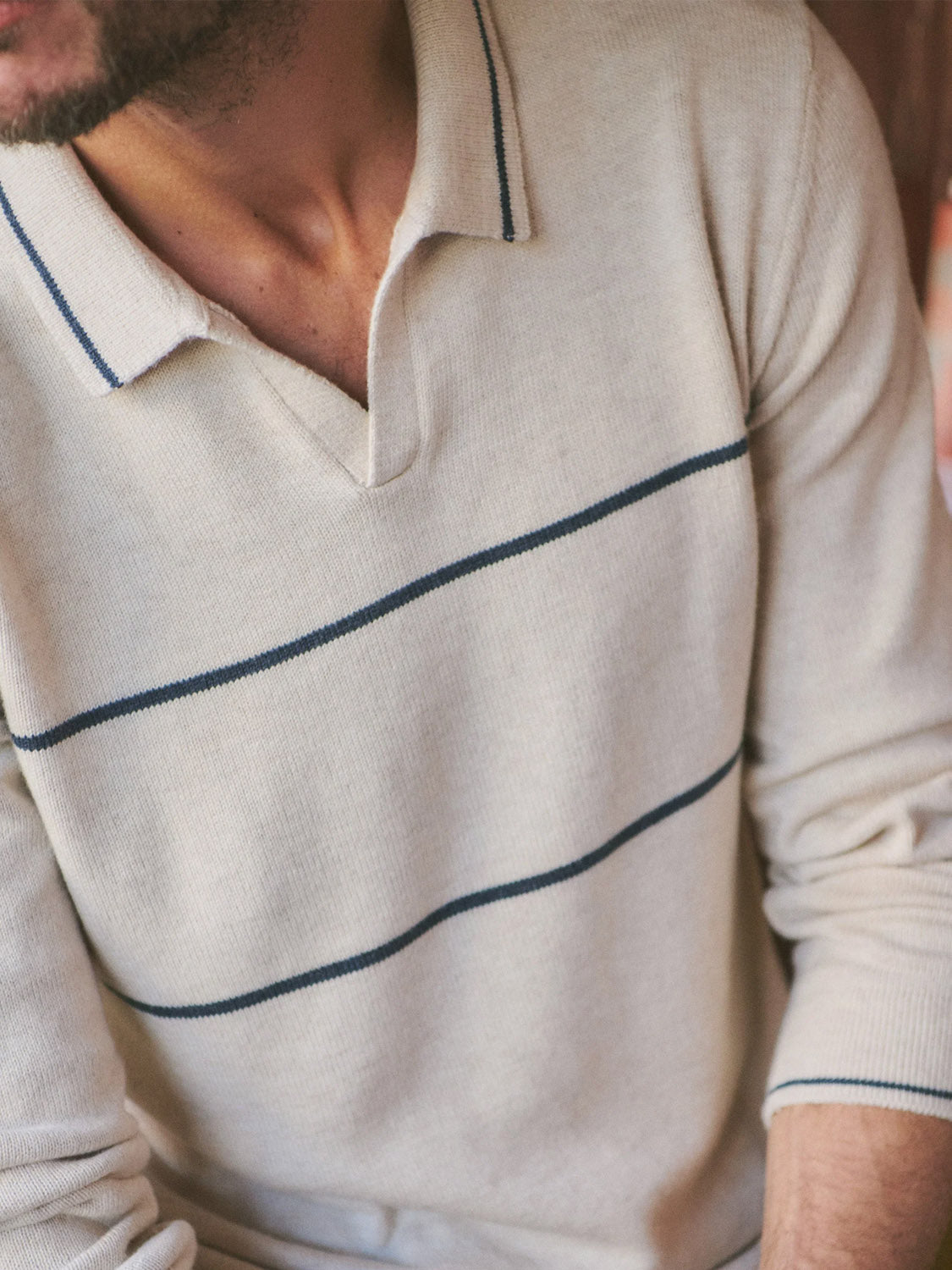 A person wearing the Marine Layer Liam Long Sleeve Sweater Polo, a cream knit shirt with a Johnny collar and thin navy blue stripes. The image is cropped to show only the torso and lower face.