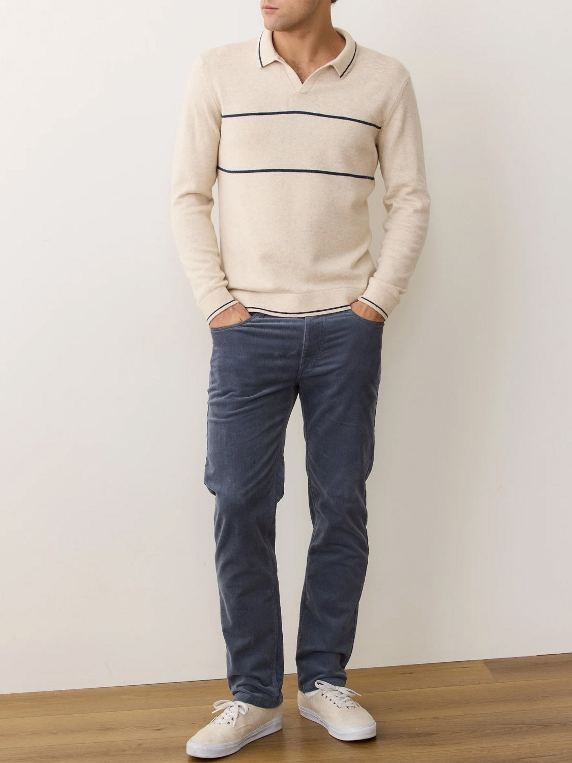 A person stands indoors against a white wall, wearing the Marine Layer Liam Long Sleeve Sweater Polo with thin horizontal stripes, blue corduroy pants, and white sneakers. Their hands are in their pockets, and their head is not visible.