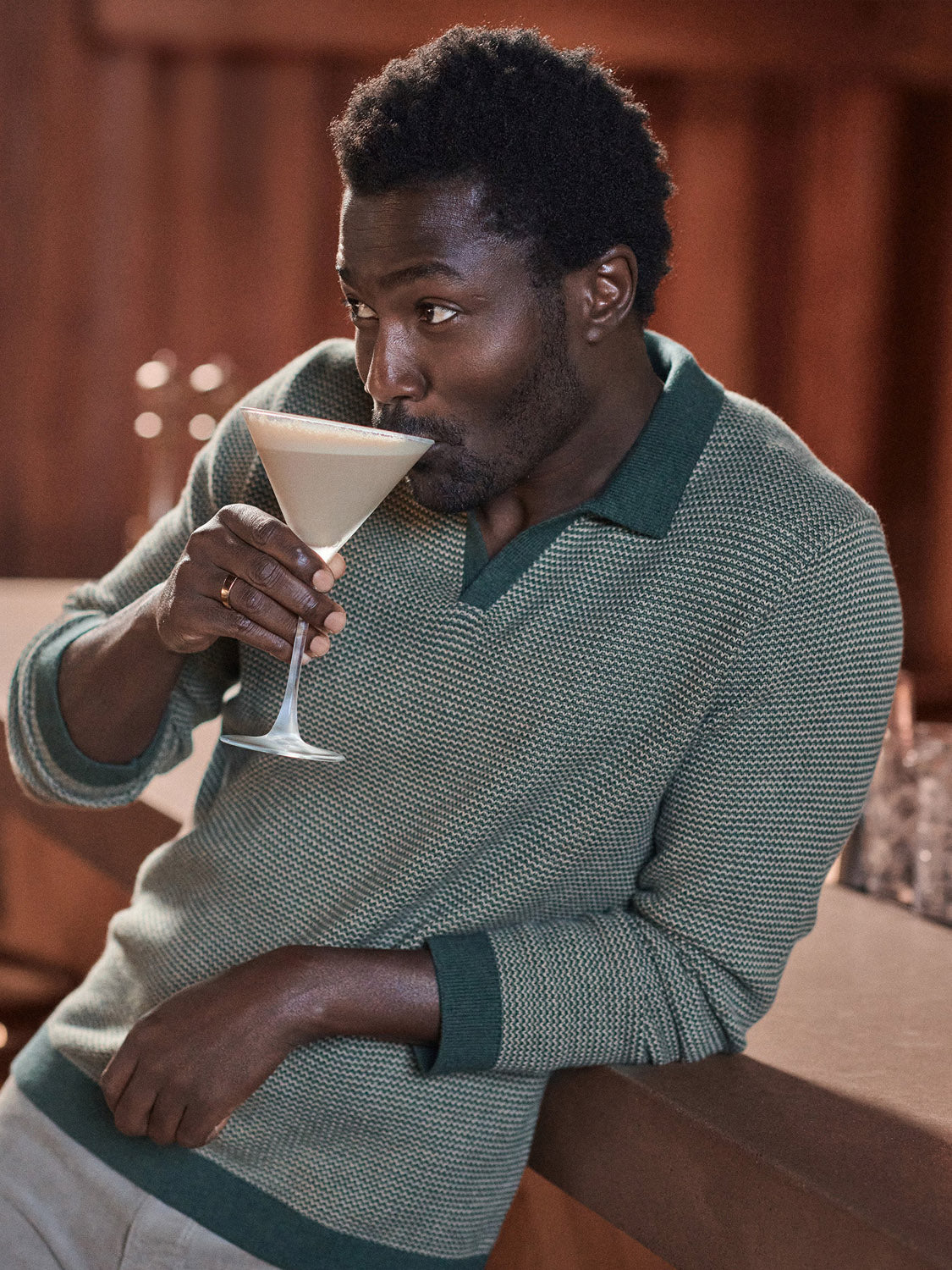 A man in the Marine Layer Liam Long Sleeve Sweater Polo in green leans against a counter, sipping a creamy cocktail from a martini glass and looking sideways with a relaxed expression.