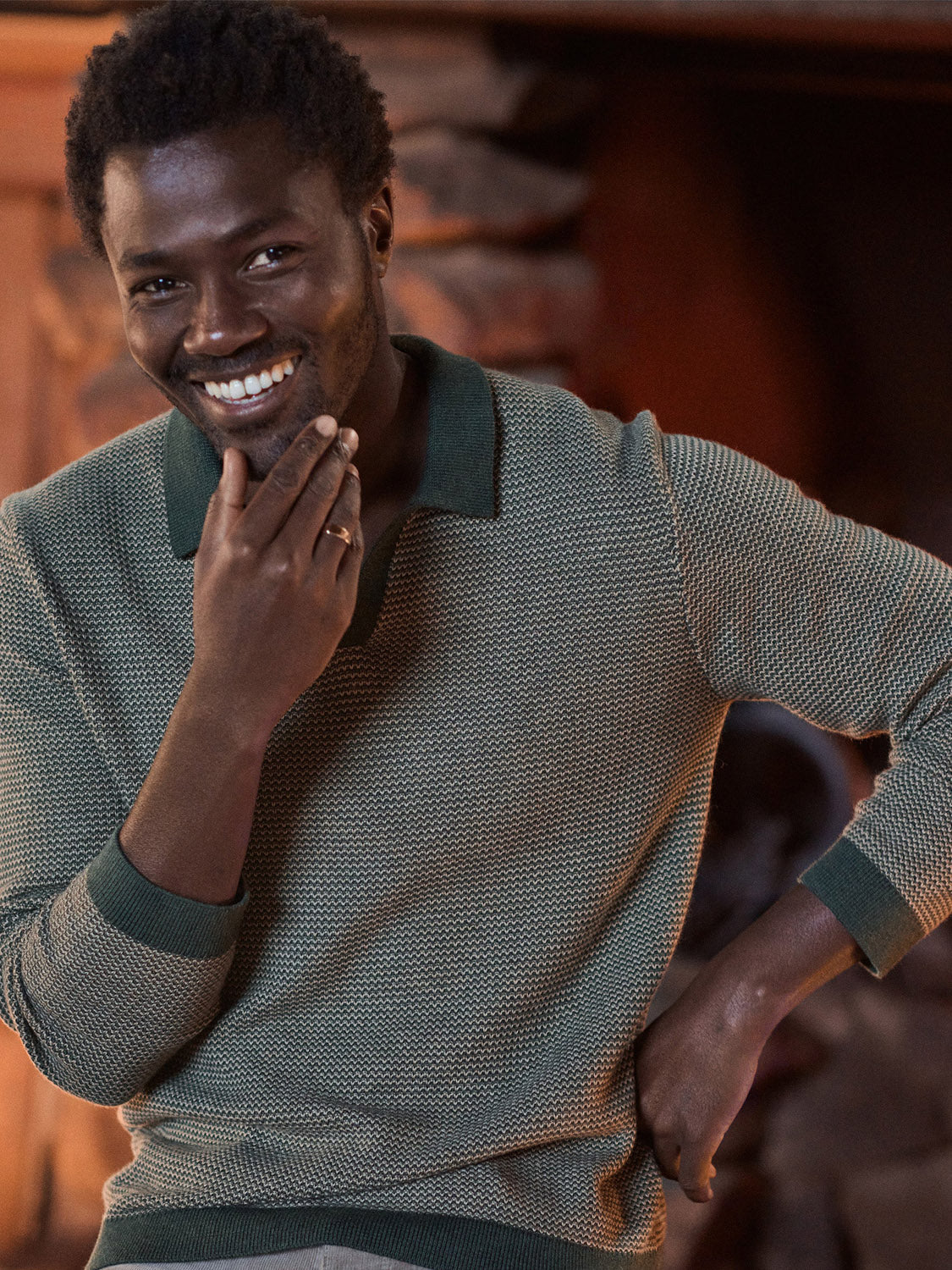 A smiling man in the Marine Layer Liam Long Sleeve Sweater Polo rests his hand on his chin and looks at the camera, set against a cozy backdrop with warm lighting and a stone fireplace.