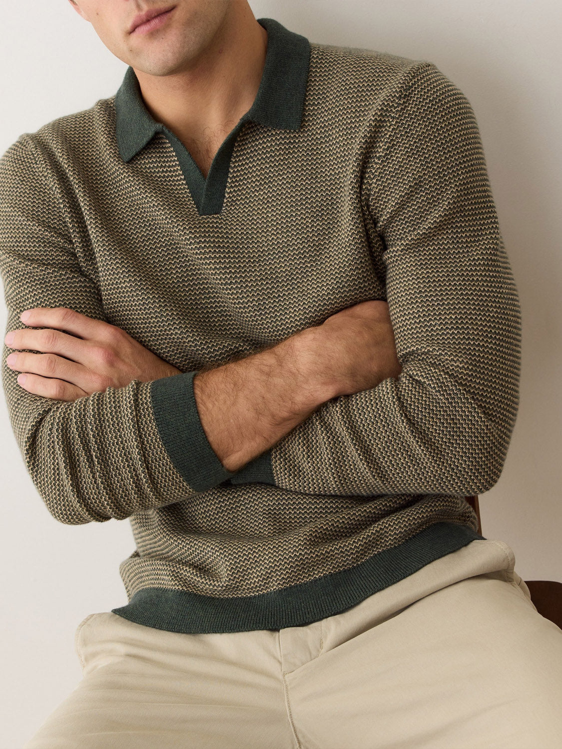 A man with light skin and short hair, partially out of frame, sits with arms crossed wearing the Marine Layer Liam Long Sleeve Sweater Polo in textured olive green, featuring a Johnny collar and dark green cuffs, paired with light beige pants.