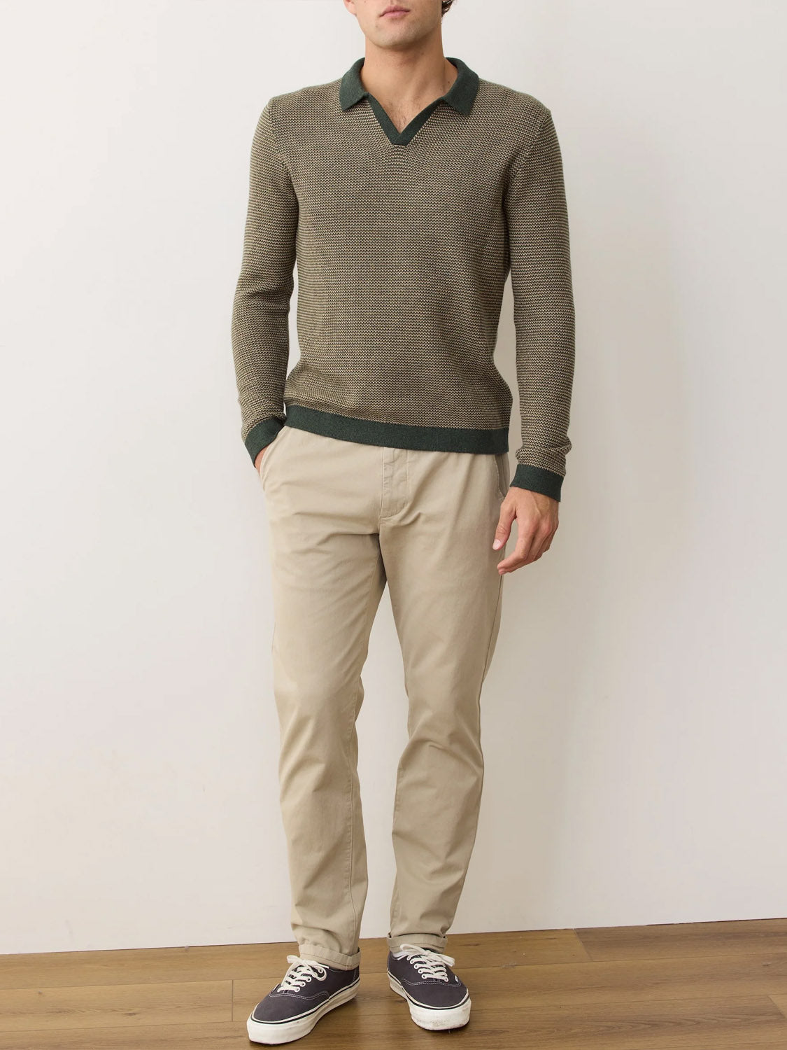 A person stands indoors against a plain wall, wearing the Marine Layer Liam Long Sleeve Sweater Polo in greenish-brown, beige pants, and dark sneakers with white soles. Their hands are in their pockets.