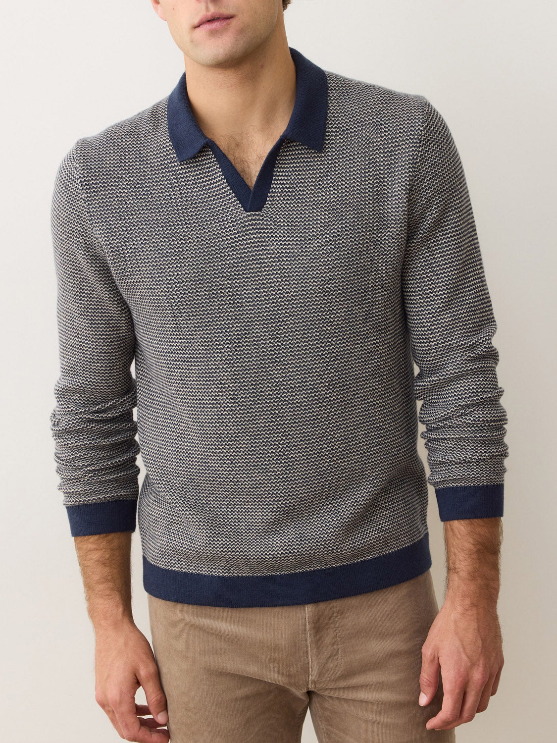 A man stands against a plain light background wearing the Marine Layer Liam Long Sleeve Sweater Polo—a textured, gray Merino wool sweater with a dark blue Johnny collar and cuffs—paired with light brown pants. His face is partially out of frame.