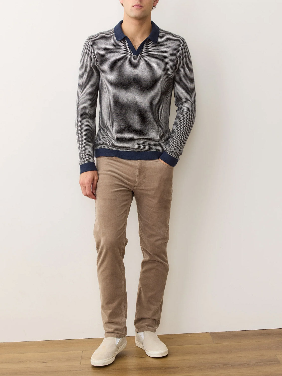 A man stands indoors against a white wall, wearing the Marine Layer Liam Long Sleeve Sweater Polo in gray, paired with beige corduroy pants and white slip-on shoes. His arms relax at his sides and his face is not shown.