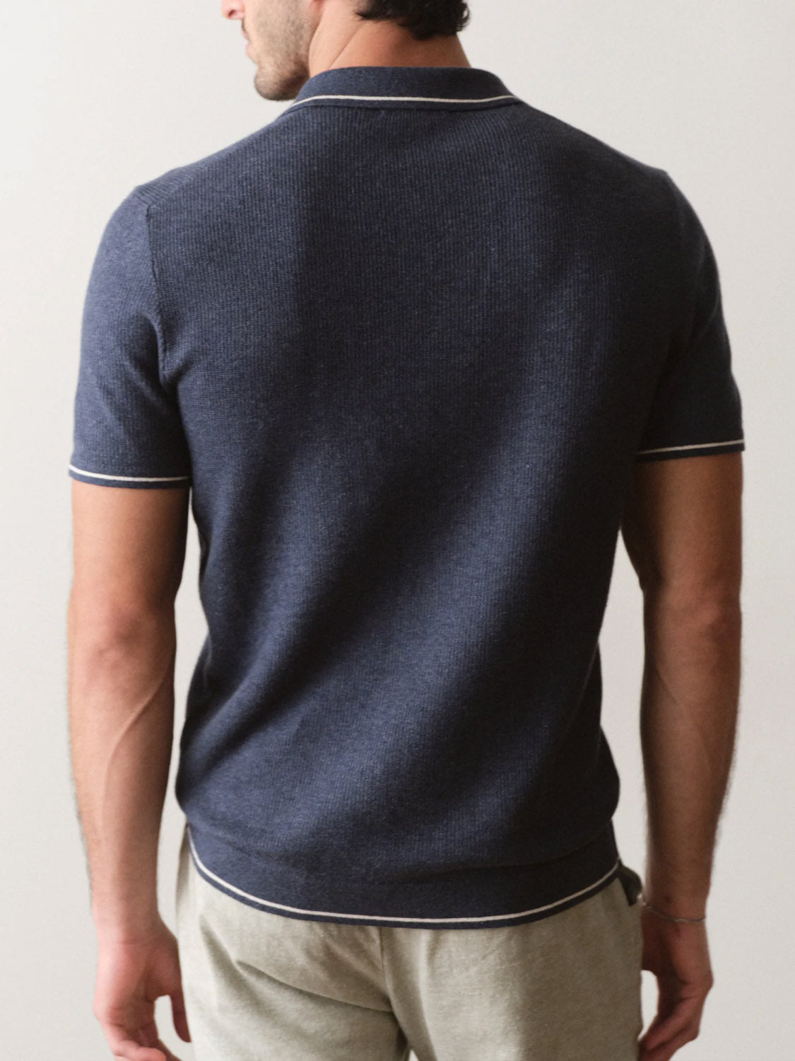 A man faces away from the camera wearing the Marine Layer Liam Sweater Short Sleeve Polo in dark navy with white trim, paired with light beige pants.