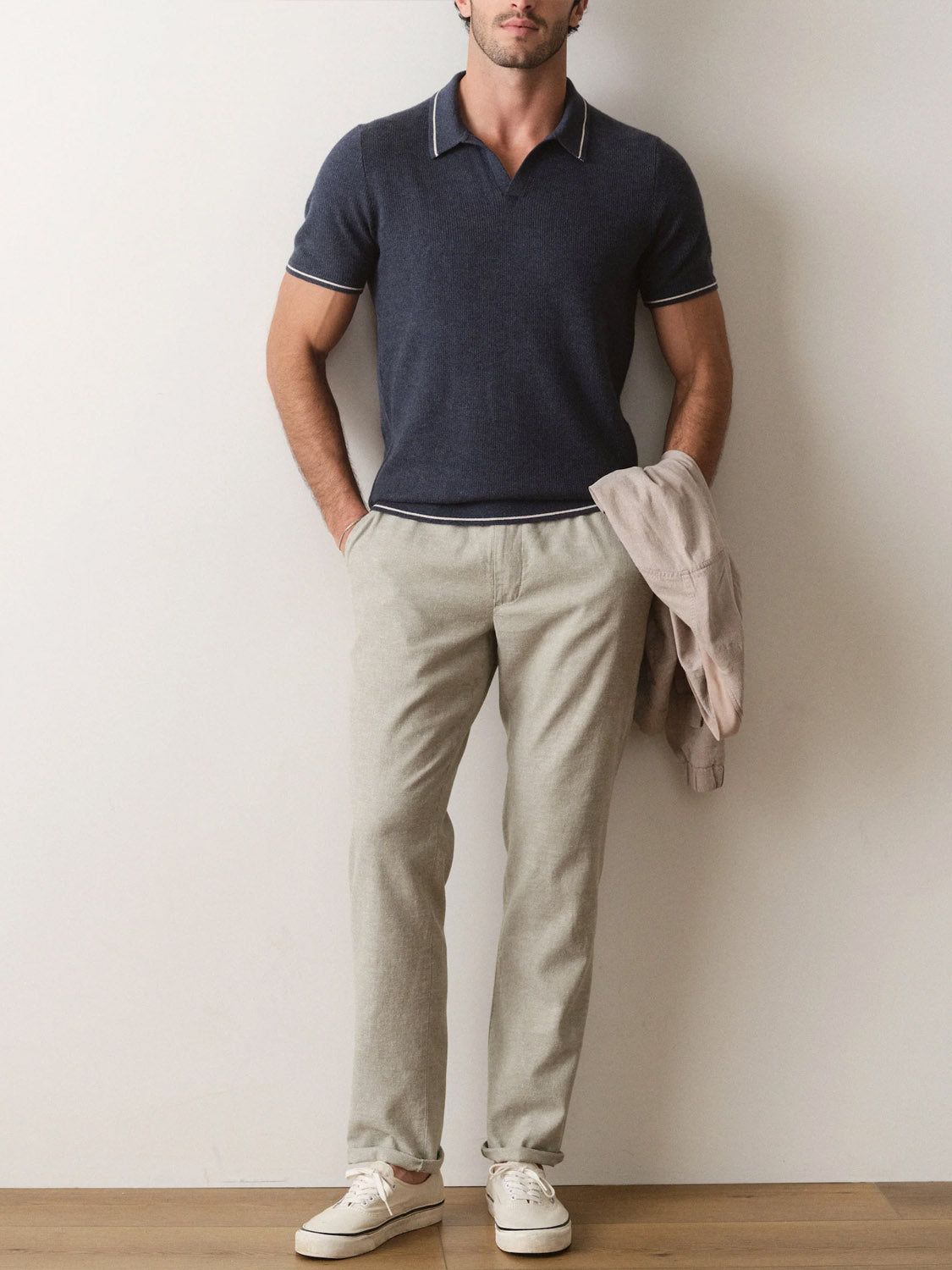 A man stands against a plain wall wearing the Marine Layer Liam Sweater Short Sleeve Polo in navy, light gray trousers, and white sneakers. He holds a beige jacket in his left hand and has his right hand in his pocket.