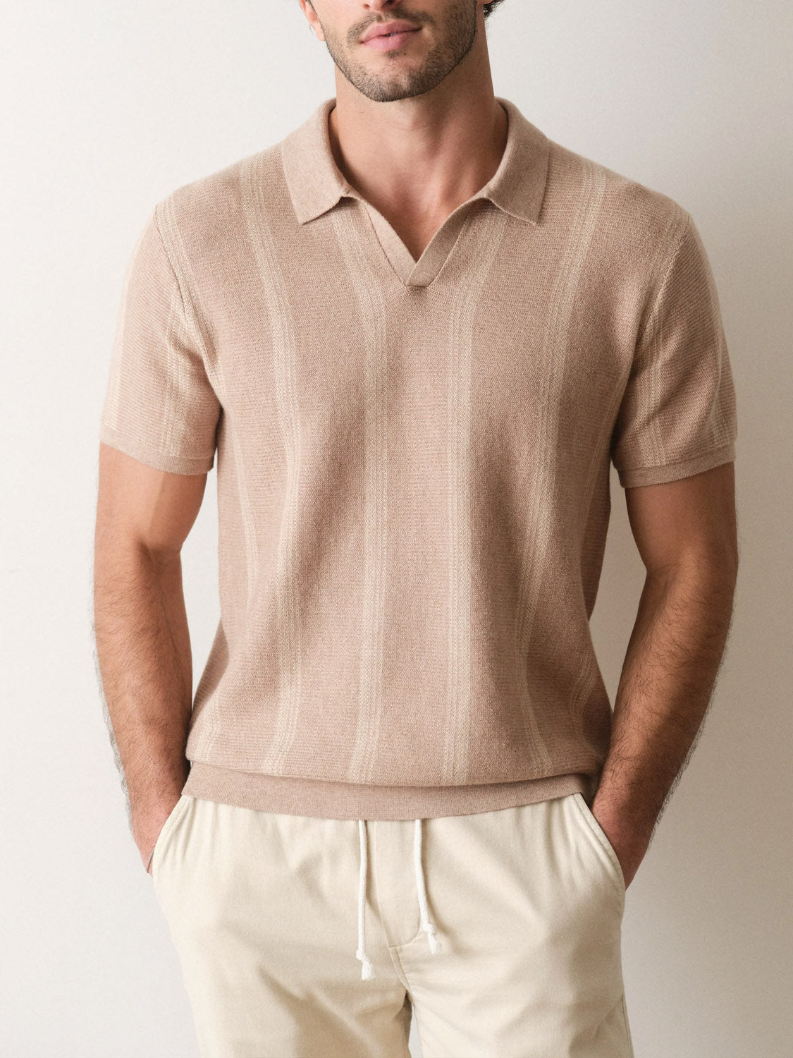 A man stands with his hands in his pockets against a plain light background, wearing the Marine Layer Liam Sweater Short Sleeve Polo and off-white drawstring pants.