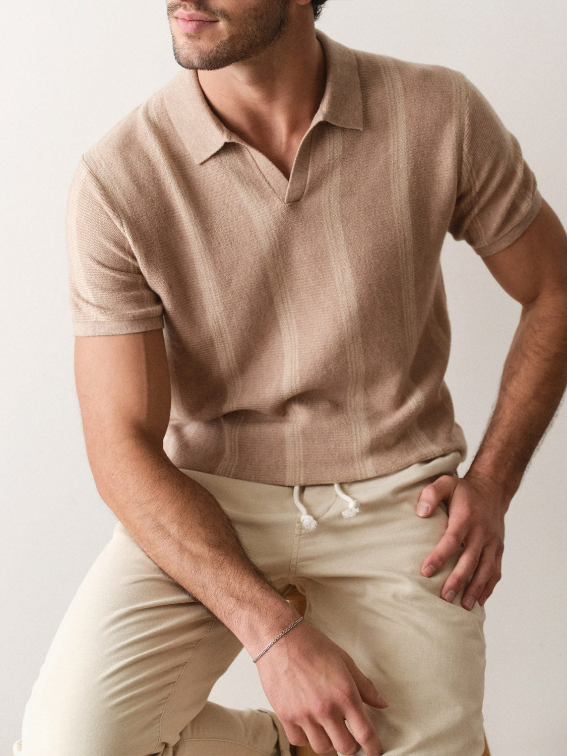 A man poses seated on a stool, wearing the Marine Layer Liam Sweater Short Sleeve Polo and beige drawstring pants. One hand rests on his knee, the other on his thigh, with his face partially visible.