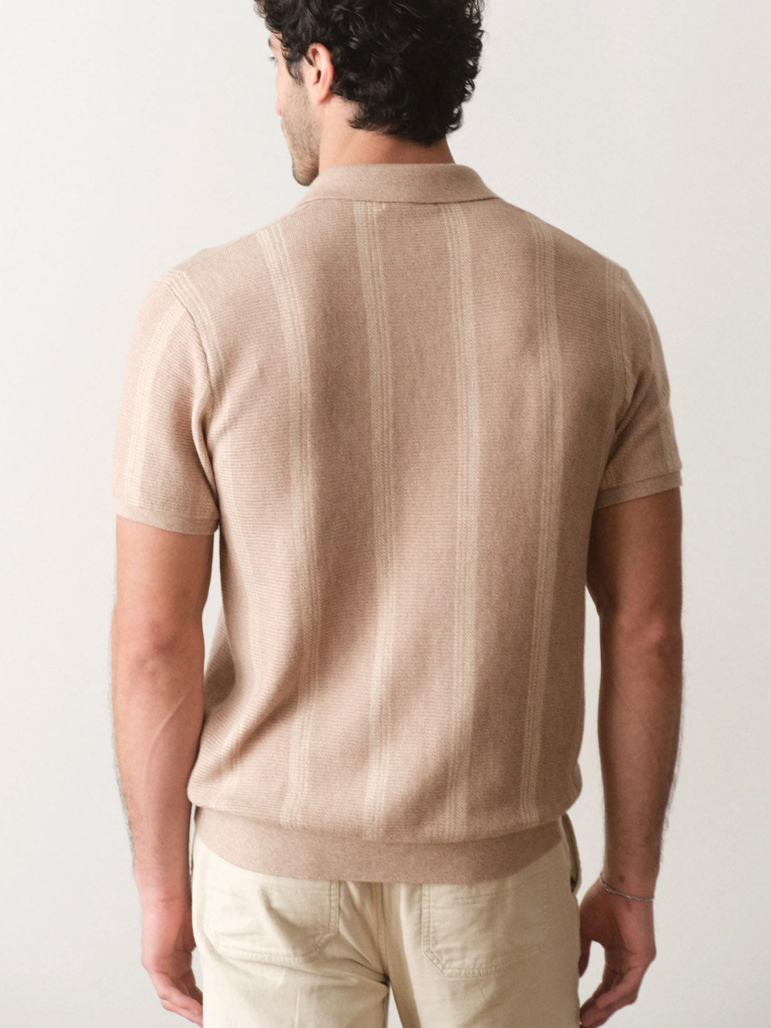 A man faces away from the camera, wearing the Marine Layer Liam Sweater Short Sleeve Polo in light brown with subtle vertical stripes and beige pants against a plain light background.