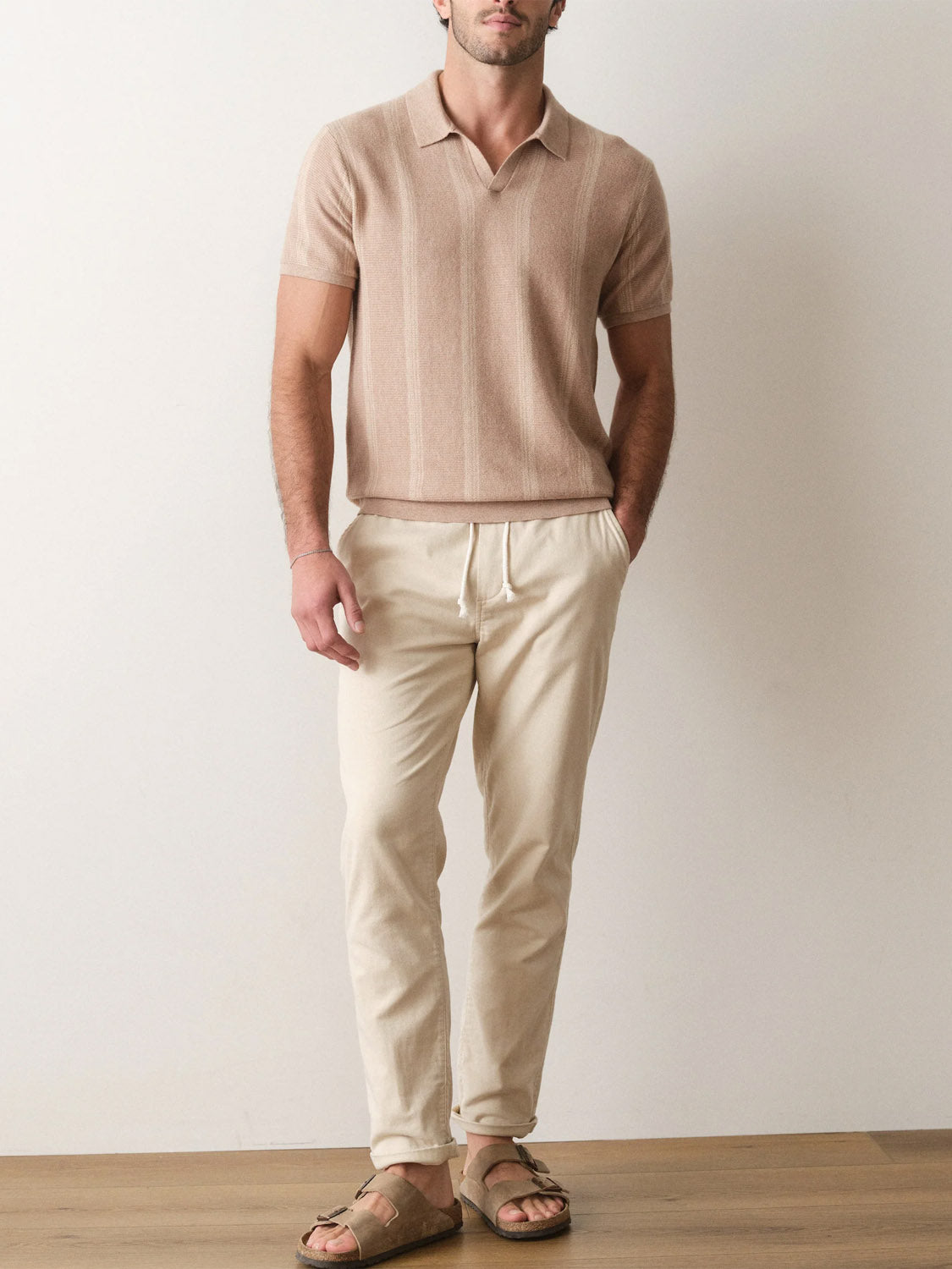 A man stands indoors against a plain wall, wearing the Marine Layer Liam Sweater Short Sleeve Polo, cream drawstring pants, and brown sandals. His hands are in his pockets and his head is slightly turned.