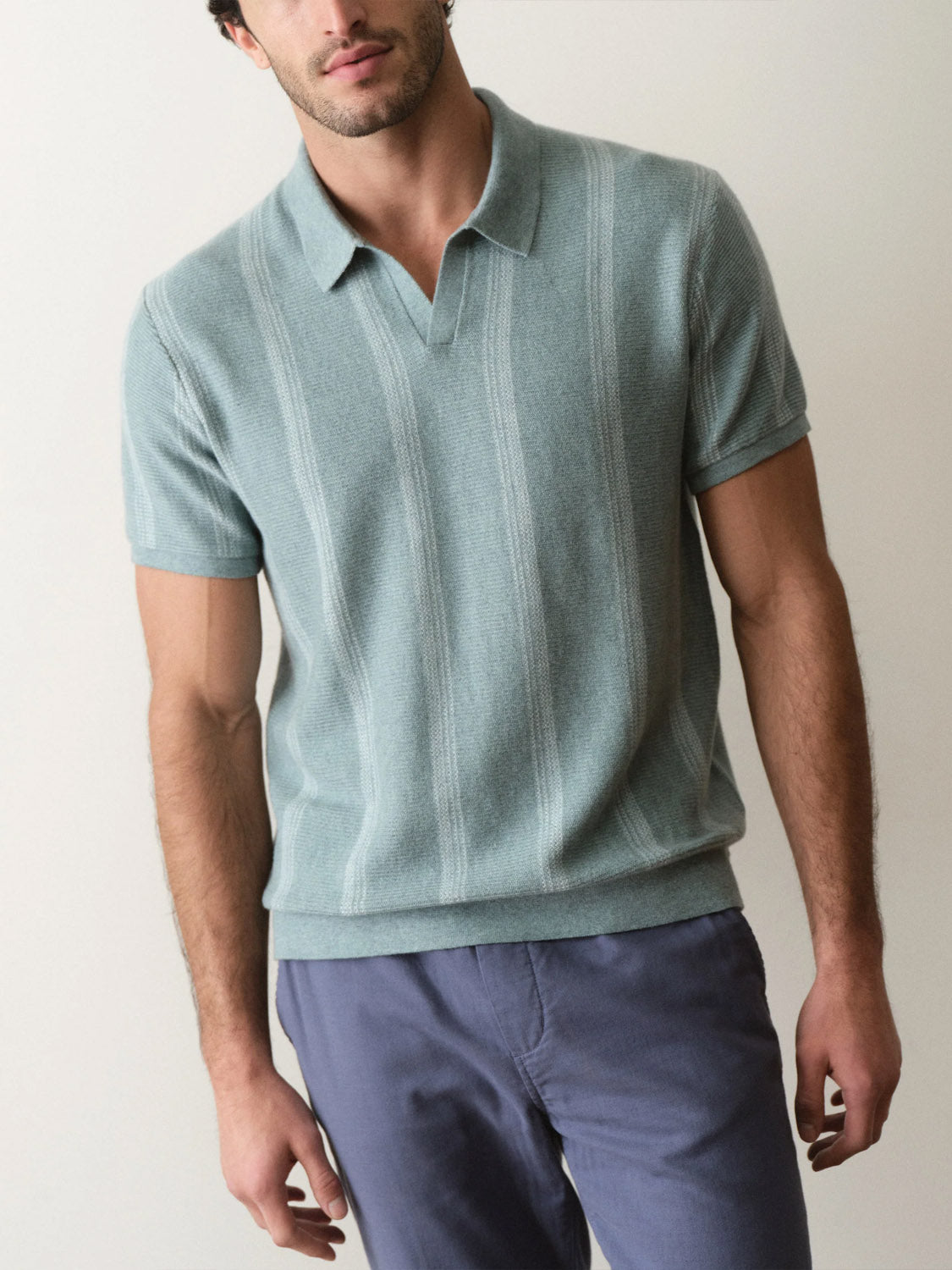 A man stands against a light background in the Marine Layer Liam Sweater Short Sleeve Polo—a light blue knit polo with subtle vertical stripes—and blue pants. His face is partially visible.