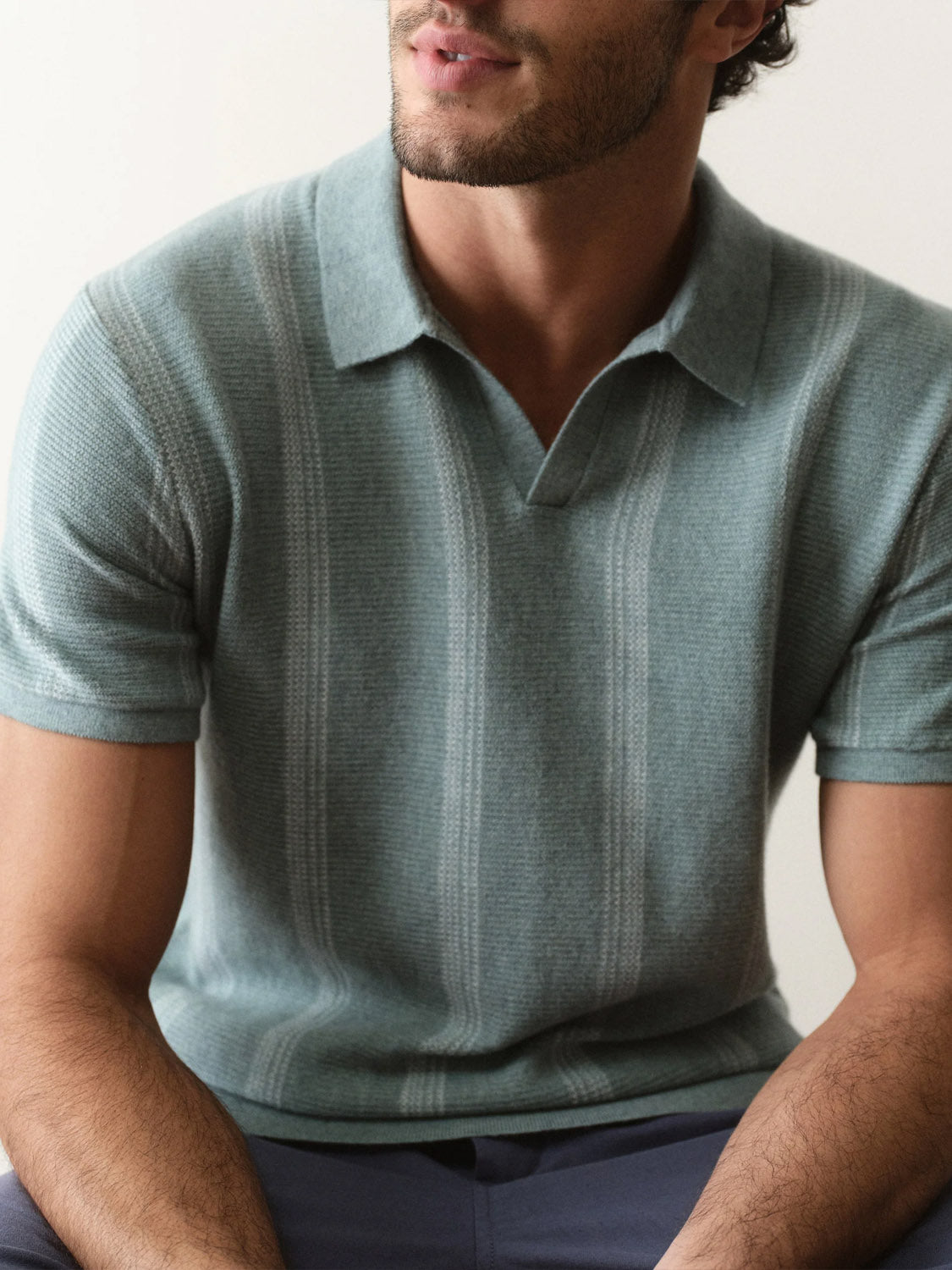 A man with short dark hair, wearing the Marine Layer Liam Sweater Short Sleeve Polo in light blue-green with subtle vertical stripes, sits with arms on his lap, facing slightly left.