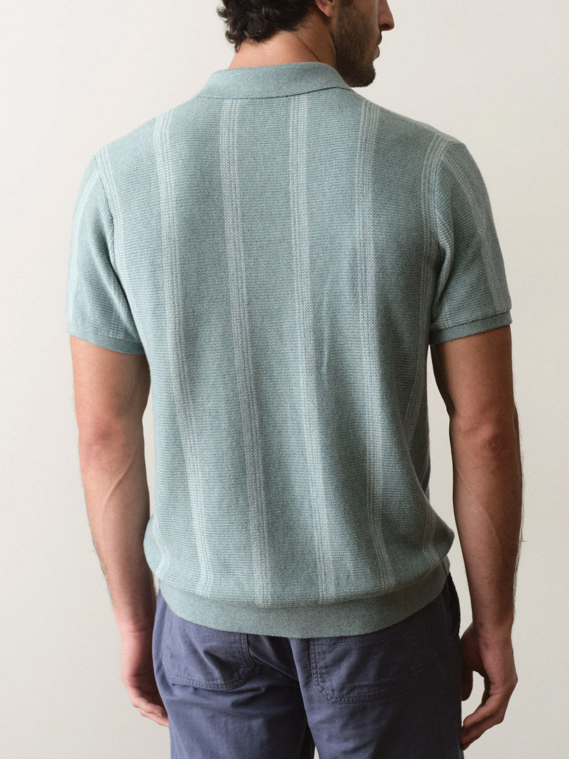 A man faces away from the camera, wearing the Marine Layer Liam Sweater Short Sleeve Polo in light green with subtle vertical stripes and a collar, paired with blue pants. The background is plain and light-colored.