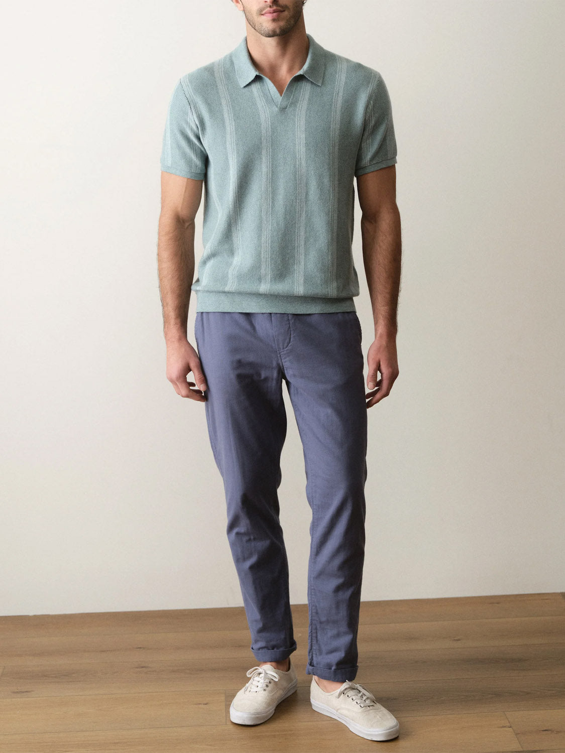 A man stands indoors on a wooden floor wearing the Marine Layer Liam Sweater Short Sleeve Polo, blue chinos, and white sneakers. His arms are relaxed at his sides as the plain background highlights his modern polo style.