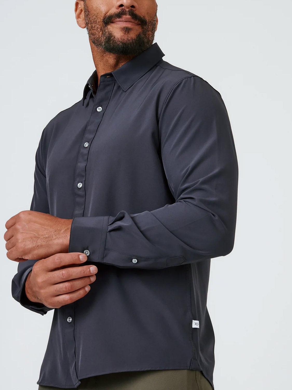 A man wears the Liberty 4-Way Stretch Shirt by ID Menswear, a dark, long-sleeve, collared button-up. Posed against a plain light background, he adjusts one sleeve with his other hand, with only part of his face visible.