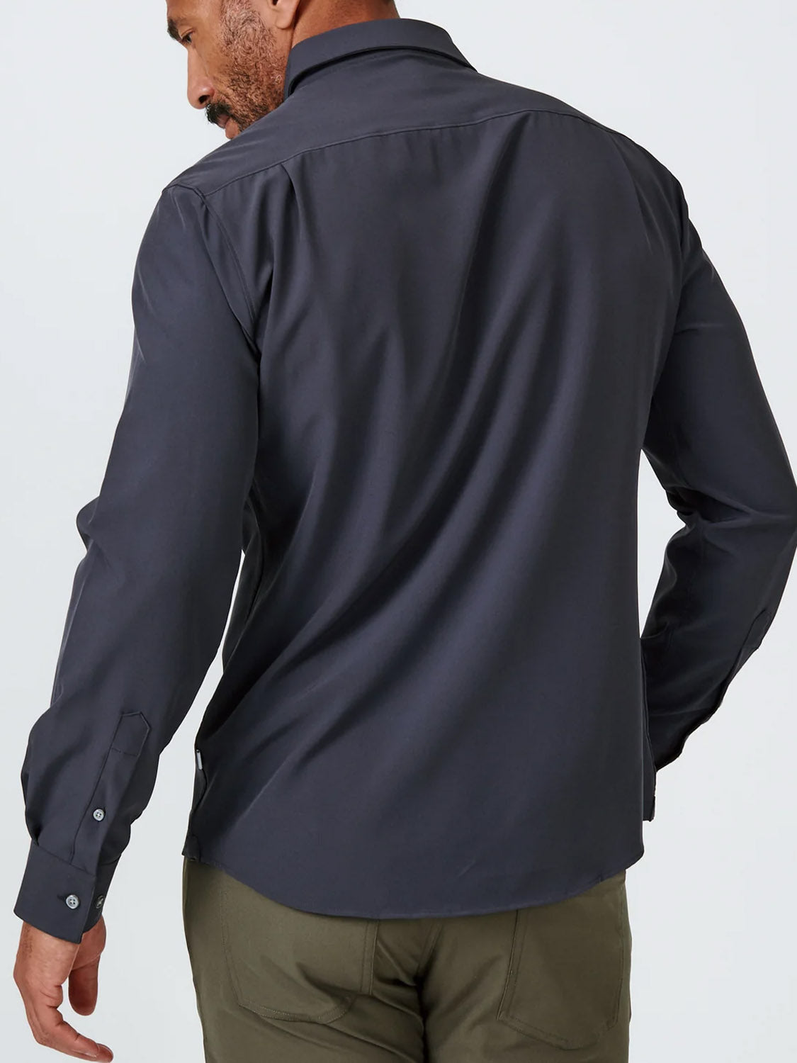 A man, seen from behind, wears the ID Menswear Liberty 4-Way Stretch Shirt in dark gray with olive green pants. He stands with his left arm slightly bent, looking over his shoulder in this stylish long-sleeve button-up.