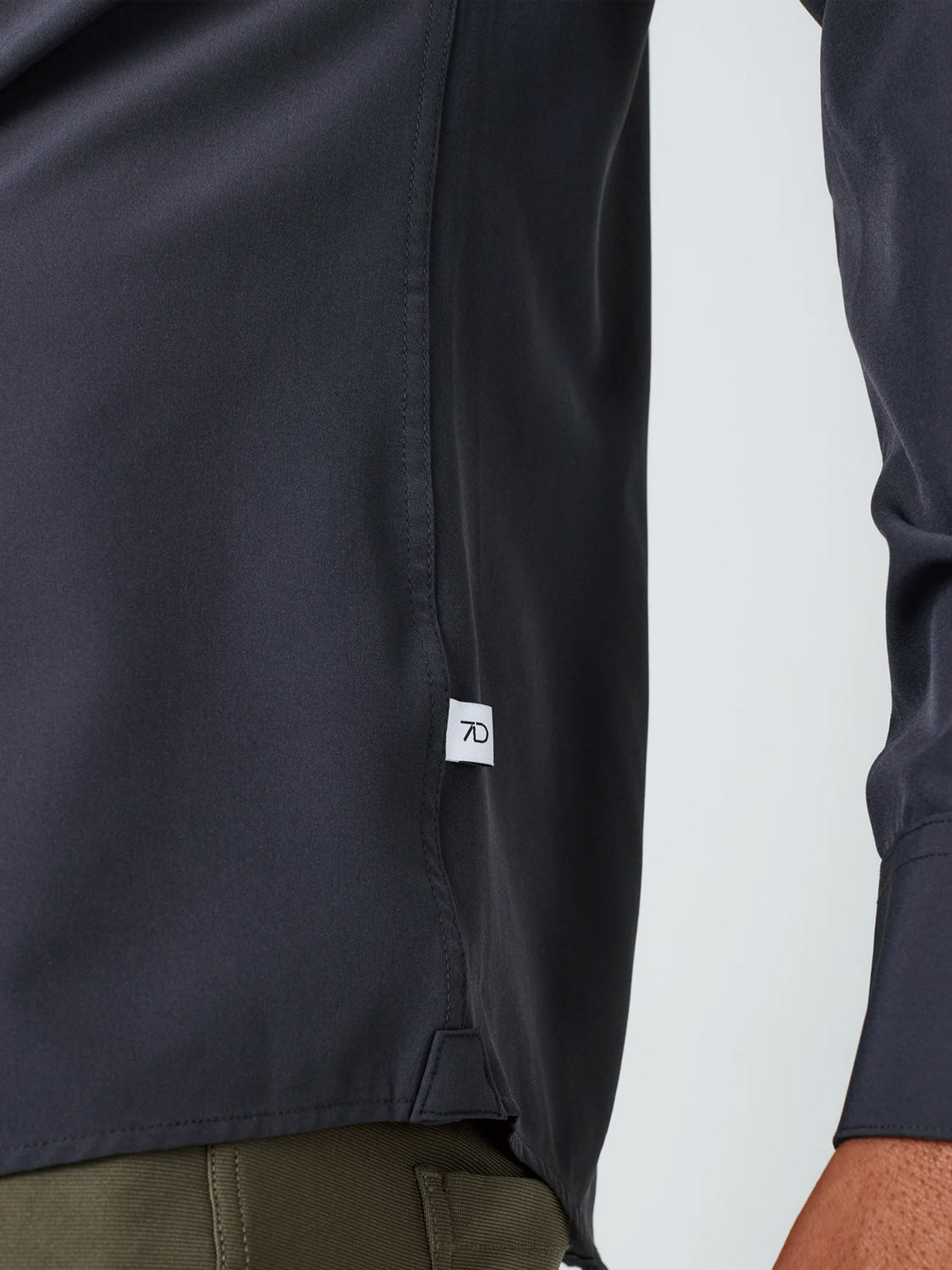 Close-up of a person’s torso wearing the Liberty 4-Way Stretch Shirt by ID Menswear in dark color, featuring a small white tag on the side seam, paired with olive green pants. Only part of the arm and hip are visible.