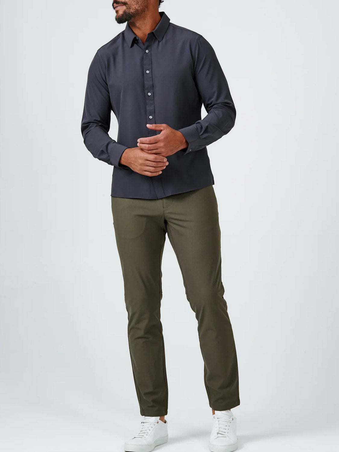 A man stands against a plain background wearing the ID Menswear Liberty 4-Way Stretch Shirt in dark grey, paired with olive green pants and white sneakers. His head is slightly turned and his hands are loosely clasped at his waist.