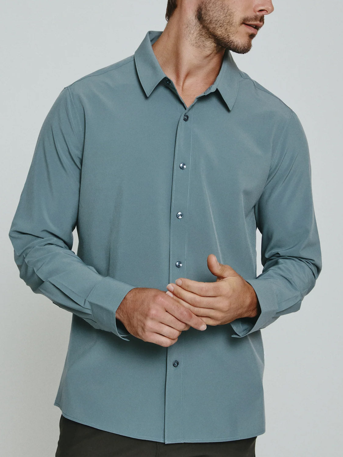 A man models the ID Menswear Liberty 4-Way Stretch Shirt, a light blue long sleeve button-up, standing against a plain background, turned slightly to the side with hands loosely clasped.