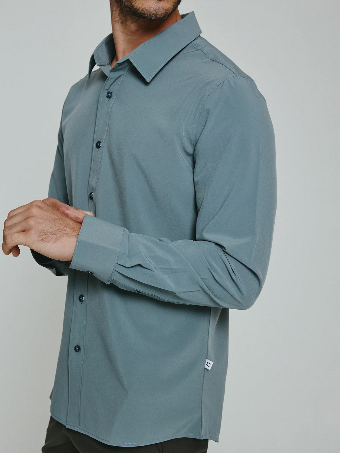 A man models the ID Menswear Liberty 4-Way Stretch Shirt, a slate-blue, long-sleeve, collared, button-up. He stands slightly side-on, head partially out of frame, hands loosely clasped in front.