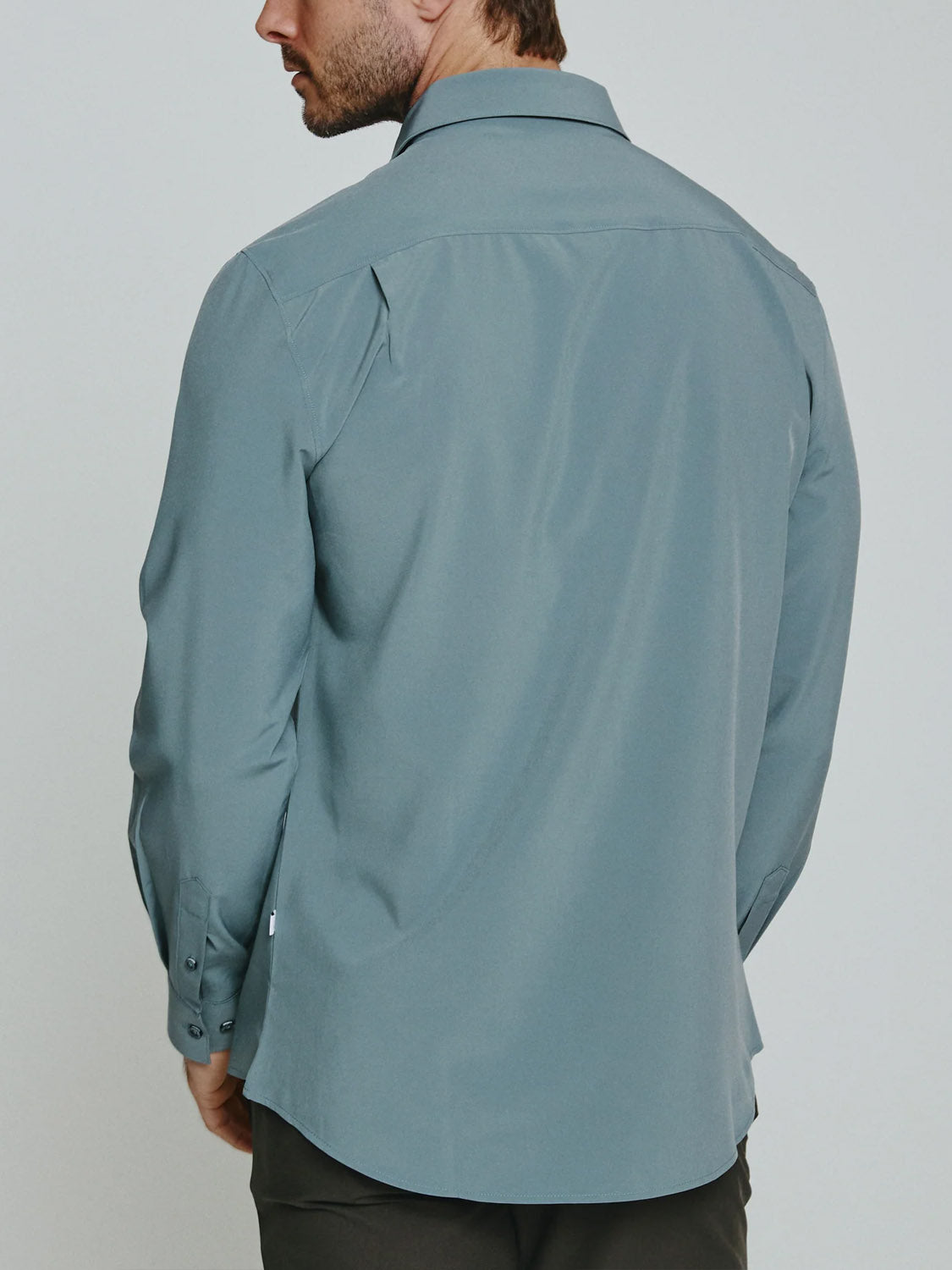 A man is shown from the back wearing the ID Menswear Liberty 4-Way Stretch Shirt in light blue, a long sleeve button-up with collar and buttoned cuffs, untucked for comfort and paired with dark pants.