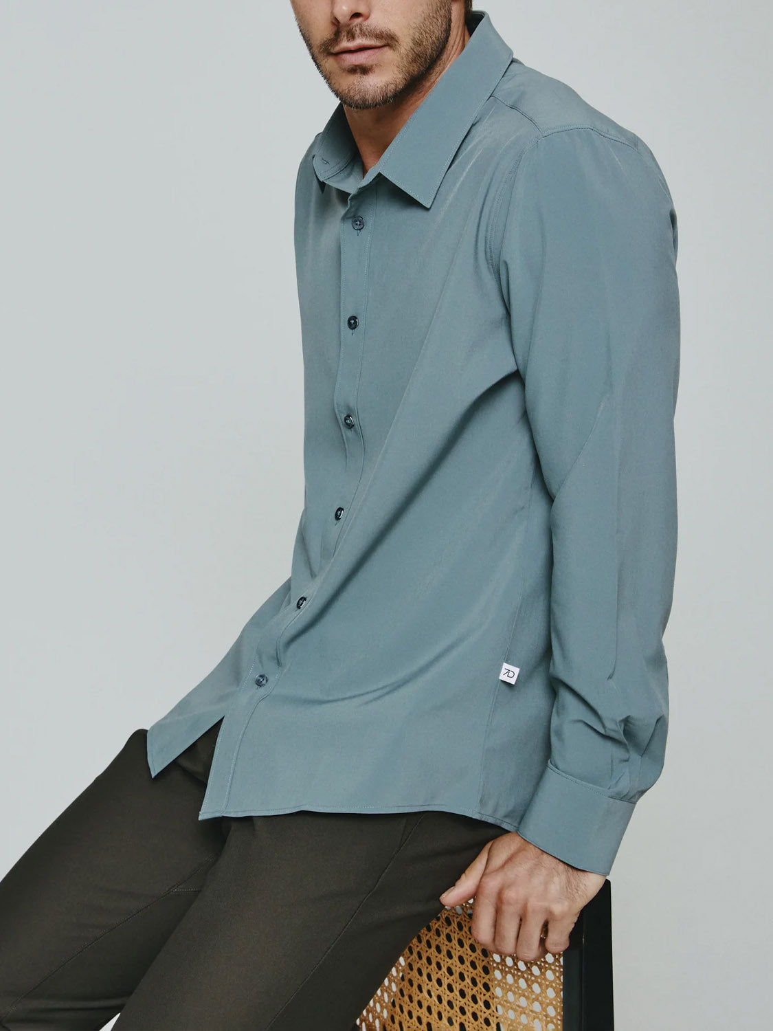 A man sits on a woven chair, wearing the Liberty 4-Way Stretch Shirt by ID Menswear in teal and dark trousers. His head is turned slightly, with his face partially out of the frame.