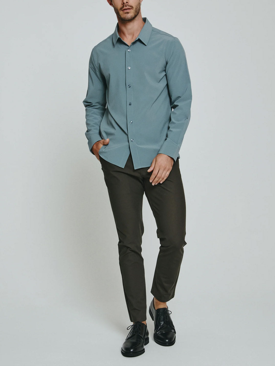 A man wearing the ID Menswear Liberty 4-Way Stretch Shirt in light blue, dark olive pants, and black dress shoes stands against a plain background with his left hand in his pocket and his right hand at his side.