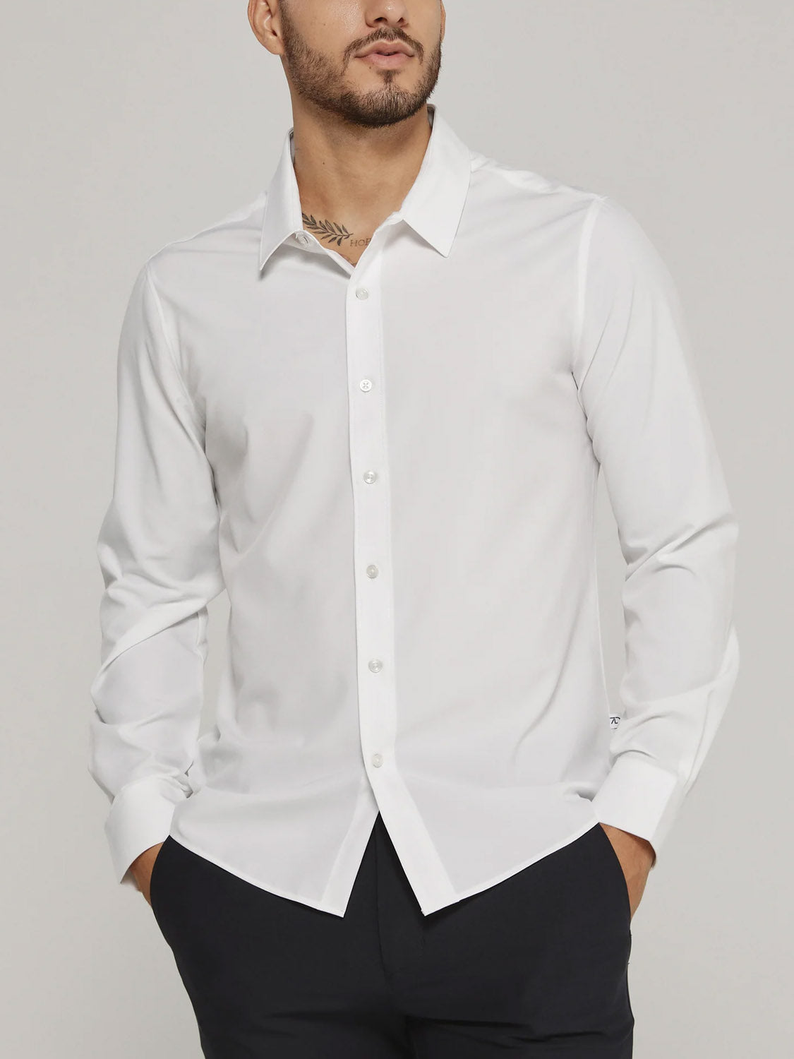 A man wearing the ID Menswear Liberty 4-Way Stretch Shirt in white with long sleeves rolled down and black pants stands with hands in pockets against a plain background.