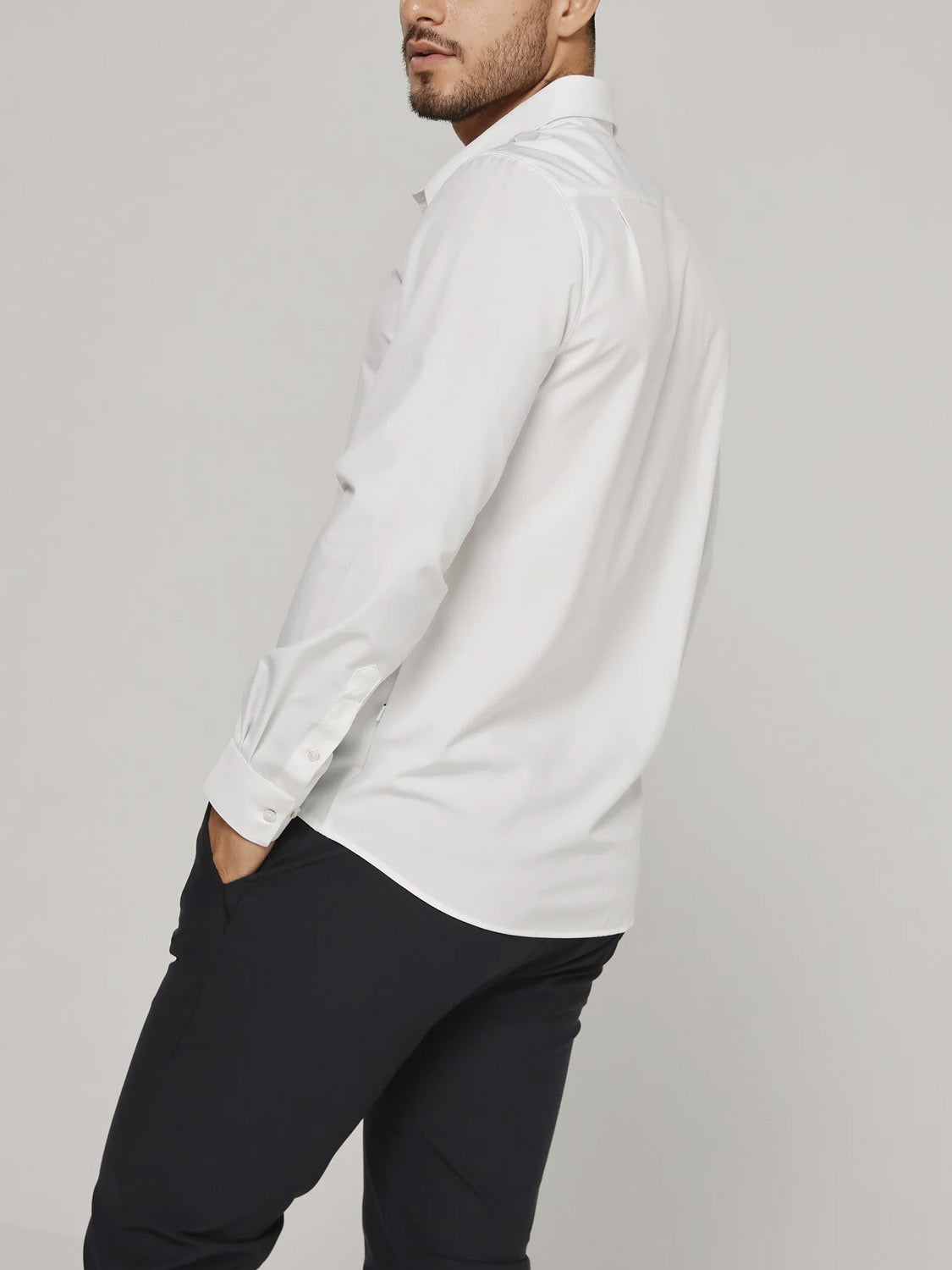 A man models the ID Menswear Liberty 4-Way Stretch Shirt in white with black pants, standing with one hand in his pocket and turned slightly from the camera against a plain light background.
