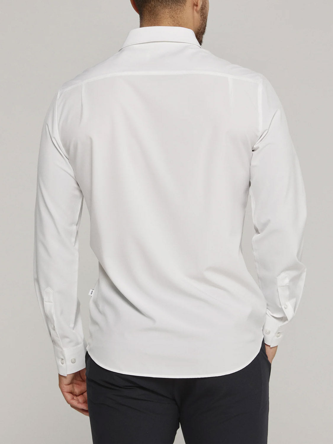 A person is shown from the back in a white Liberty 4-Way Stretch Shirt by ID Menswear with dark pants, left hand in pocket, against a plain light background.
