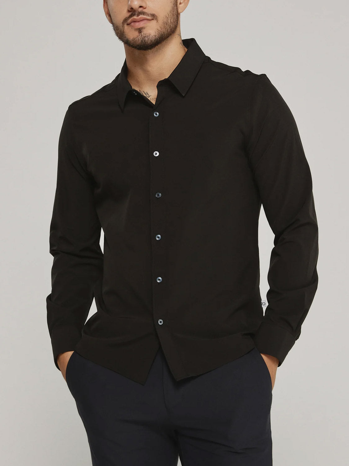 A man wears the ID Menswear Liberty 4-Way Stretch Shirt in black with sleeves down and top button undone, paired with dark pants. His hands are in his pockets, and his face is partially out of frame.
