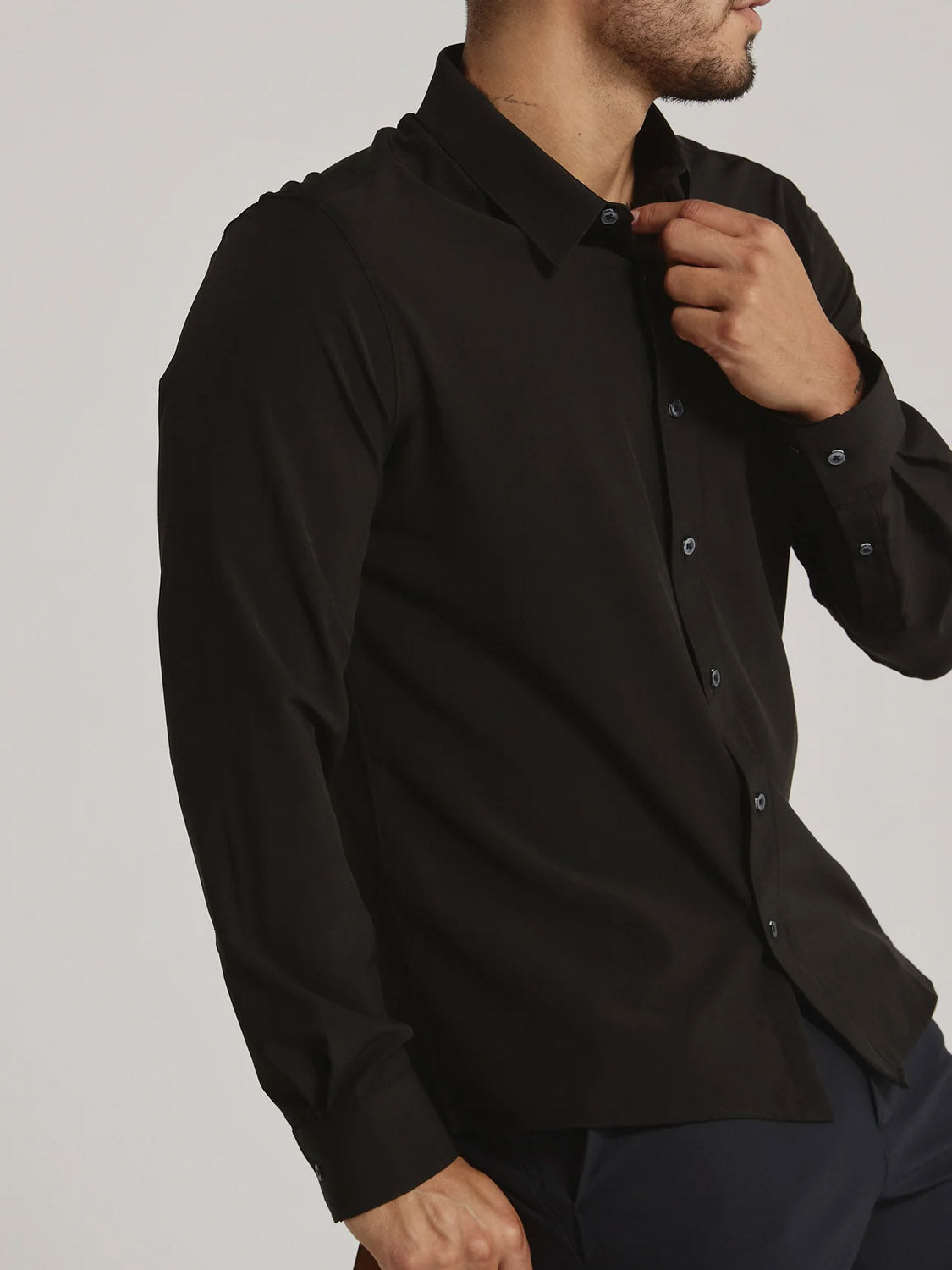 A man adjusts the top button of a fitted black Liberty 4-Way Stretch Shirt by ID Menswear. His face is partially out of frame against a plain, light-colored background.