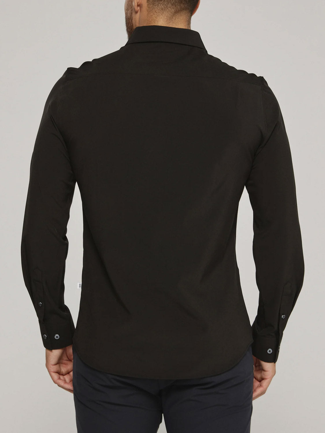 A man is seen from the back wearing the black Liberty 4-Way Stretch Shirt by ID Menswear with dark pants, standing against a plain light background. His hands rest relaxed at his sides.