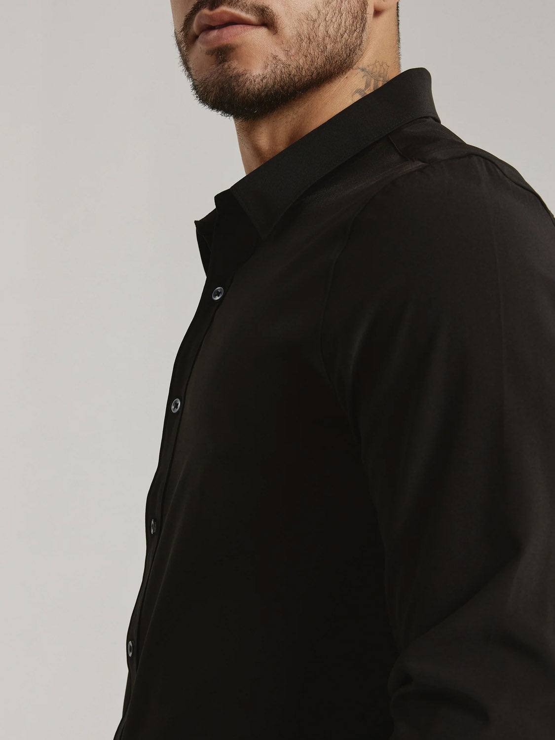 A man with a short beard is shown from the shoulders up in a fitted black ID Menswear Liberty 4-Way Stretch Shirt, seen from the side and partially from the front, against a plain light background.