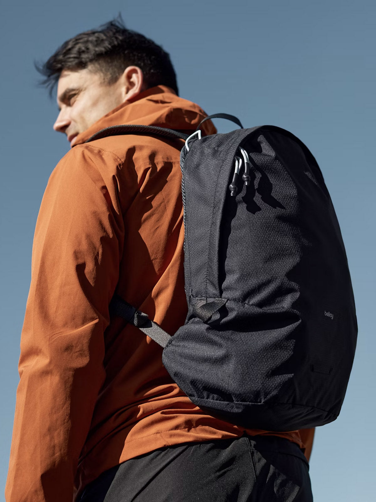 A person carries the Bellroy Lite Daypack - 20L and wears an orange jacket while standing outdoors against a clear blue sky, gazing off to the side.